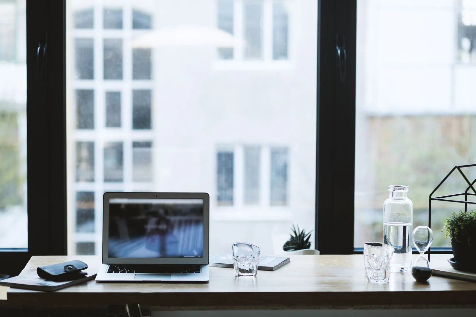 A minimal wooden desk with a laptop, a glass of water and a small plant beside a large window in soft daylight — the everyday room a workation changes